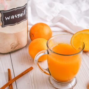 Wassail mix in the background with oranges and cinnamon sticks. A cup of hot wassail with an orange on the rim of the clear mug.