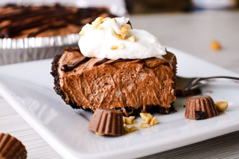peanut butter cocoa mousse pie on a plate with a bite taken out of it. It's topped with whipped cream, peanuts, and mini peanut butter cups. The pie is in the background.